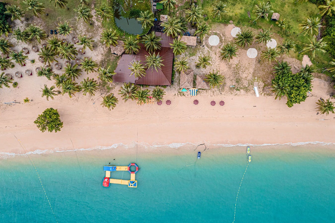 鸟瞰海滩，碧绿的海水、海滩遮阳伞、棕榈树和被绿色环绕的木制建筑。