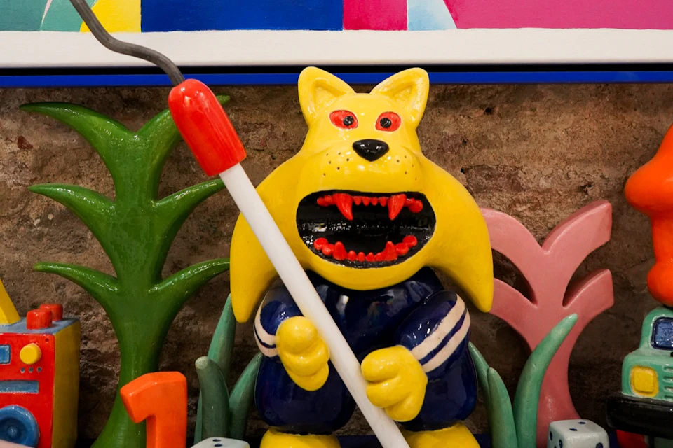 A colorful ceramic sculpture of a yellow creature with an open mouth holding a red-tipped white stick, surrounded by plants.
