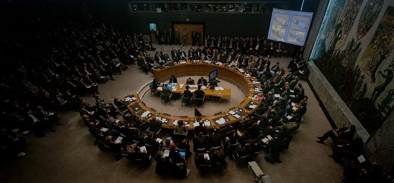 A large, circular conference table filled with people engaged in discussion, with additional attendees seated around the room.