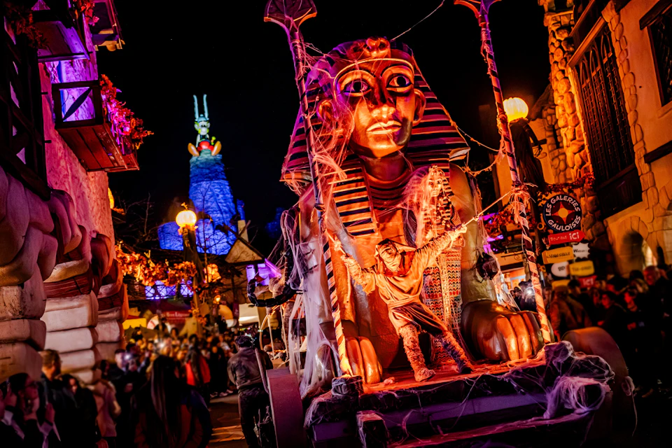 A vibrant scene featuring an illuminated Egyptian-themed float with a sphinx figure at a night-time parade or festival.