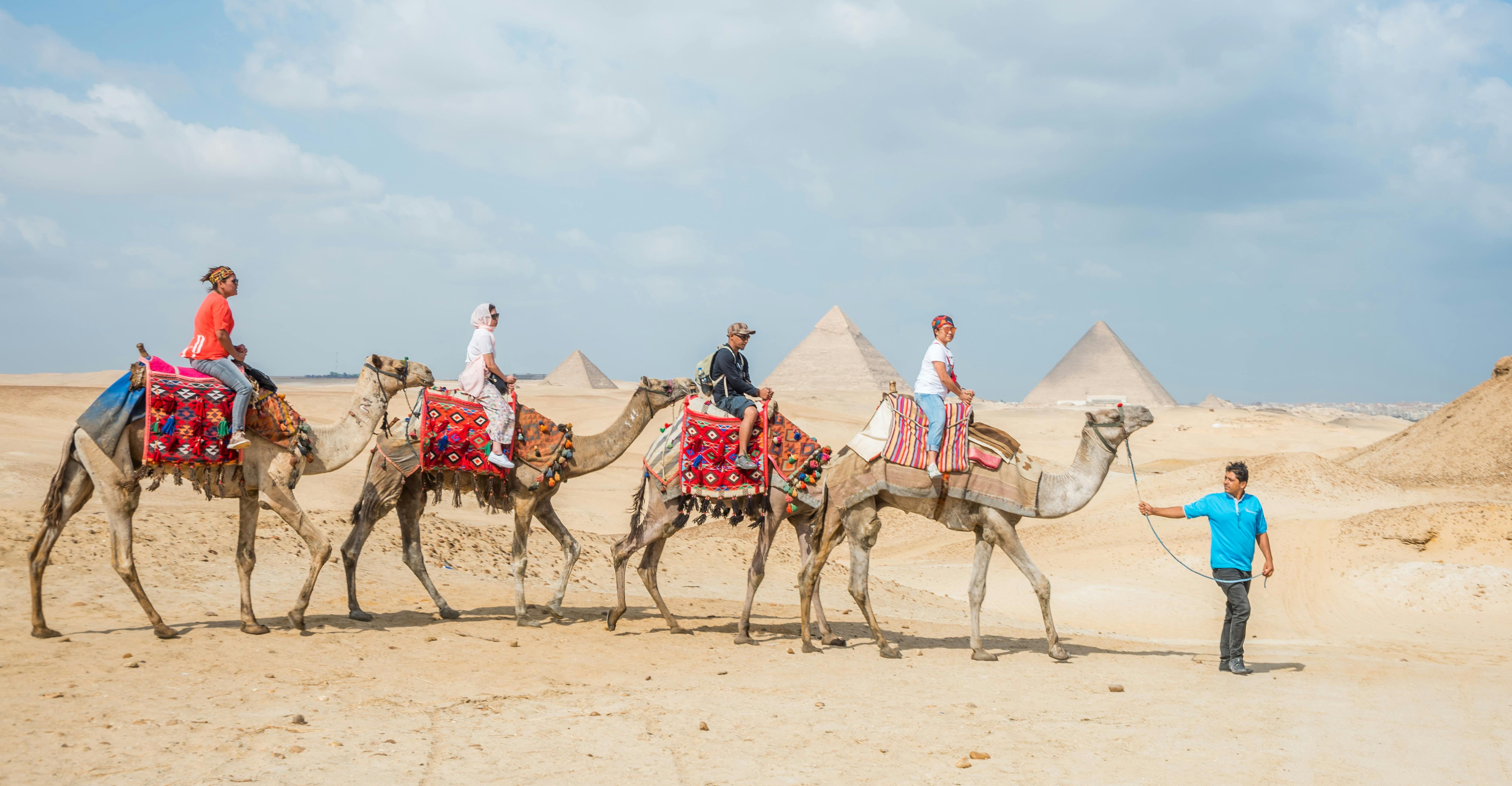 在沙漠中，三个人骑着五颜六色的马鞍骑着骆驼，背景是多云的天空下的金字塔。