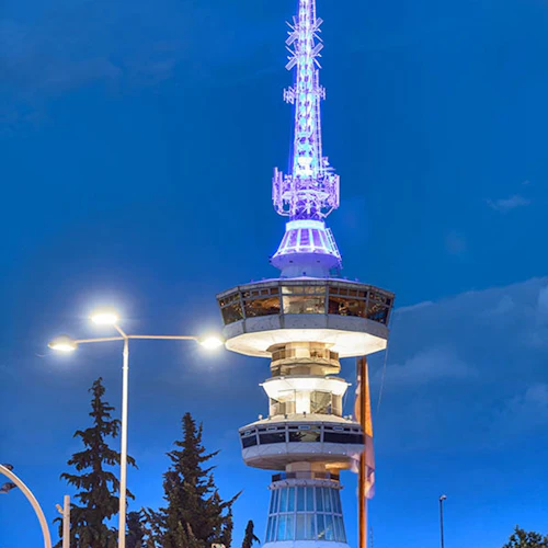 Ett högt, modernt torn upplyst med blått ljus mot en skymningshimmel, omgivet av träd och gatubelysning.