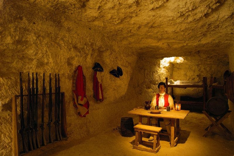 Δωμάτιο σαν σπηλιά με ένα άτομο να κάθεται σε ένα ξύλινο τραπέζι. Ρούχα και καπέλα κρέμονται στον τοίχο. Μια κουκέτα και ένα βαρέλι είναι ορατά.