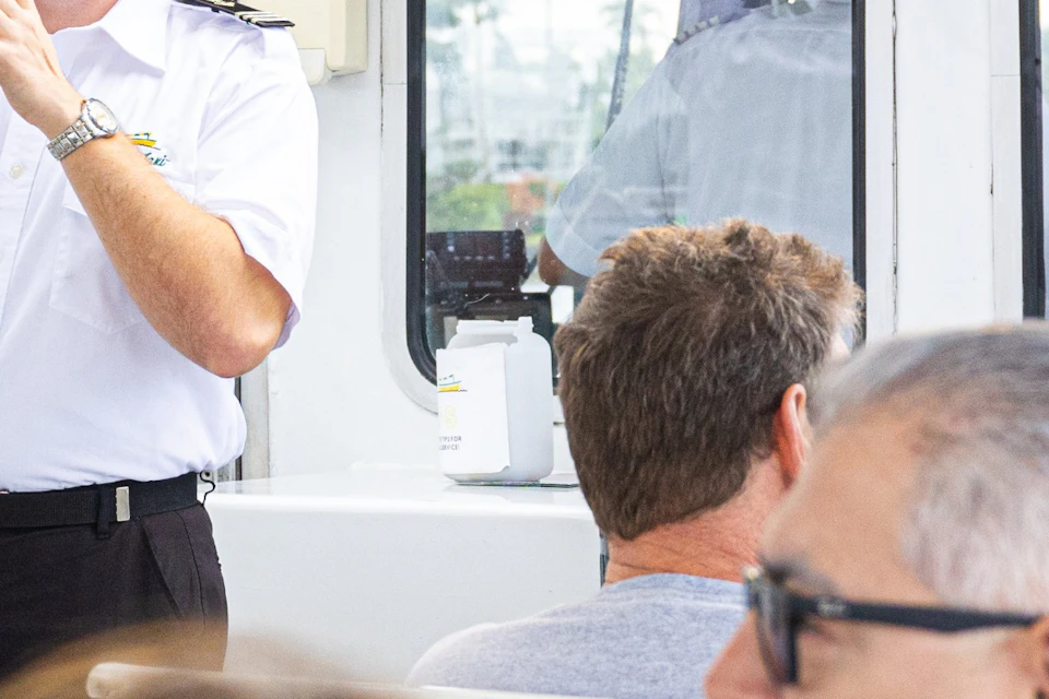 A tour guide wearing a white shirt and green cap speaks to passengers on a boat with a waterfront view in the background.