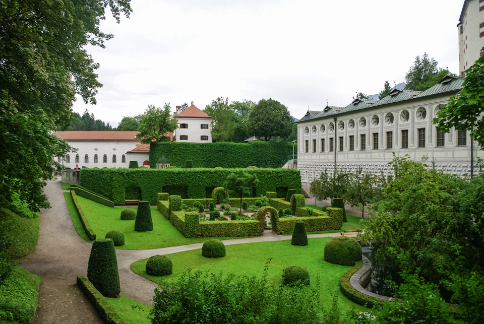 Formal garden with manicured hedges, topiaries, and pathways, adjacent to white buildings and lush green trees.