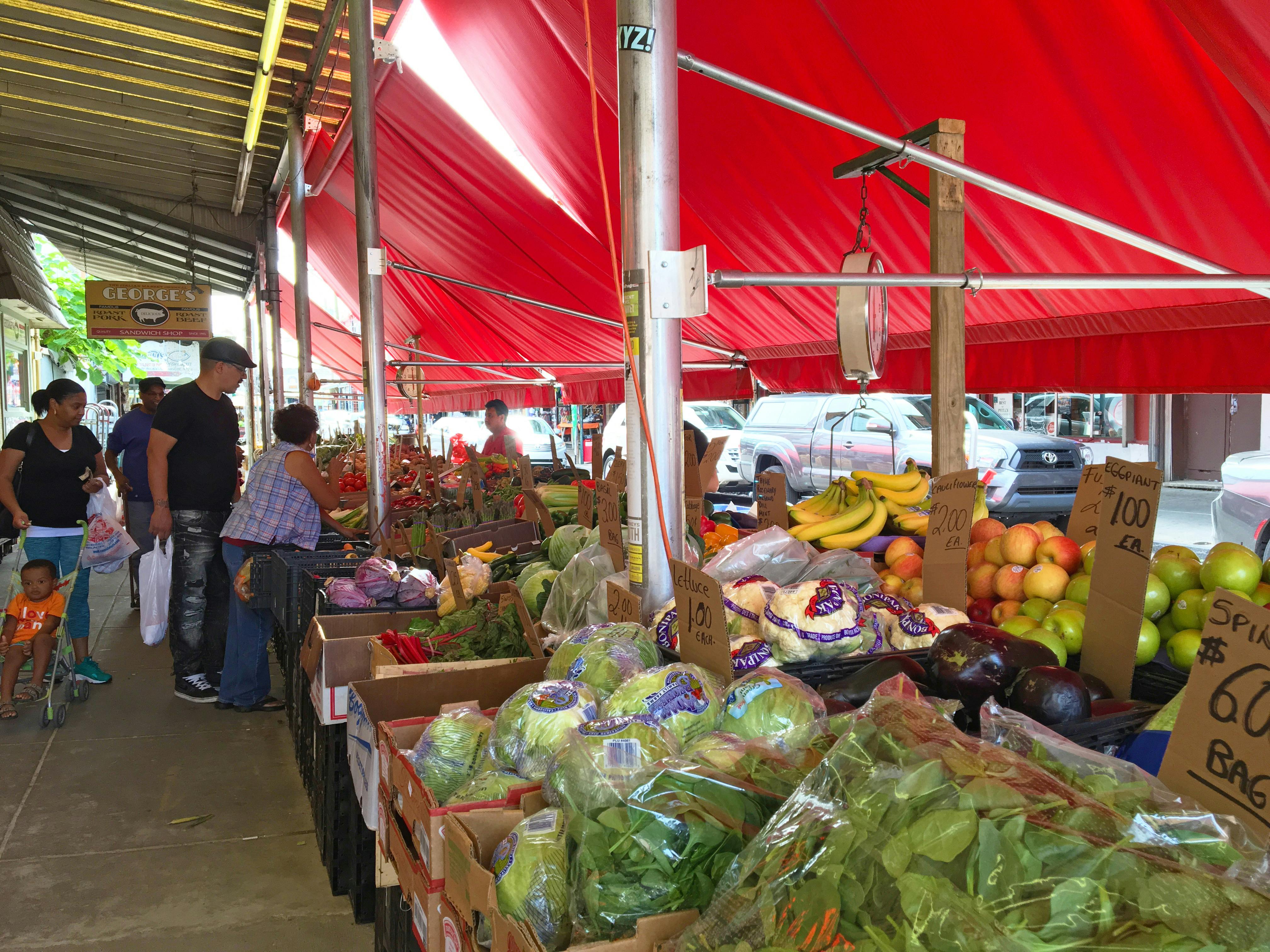 Buitenmarkt met rode luifels waar verschillende groenten en fruit worden uitgestald. Mensen snuffelen en winkelen.