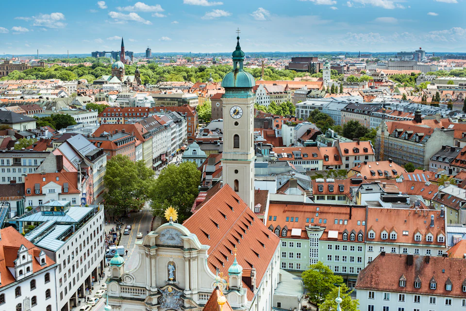 A cityscape featuring a mix of historical buildings, a prominent clock tower, and red-tiled roofs under a bright blue sky.