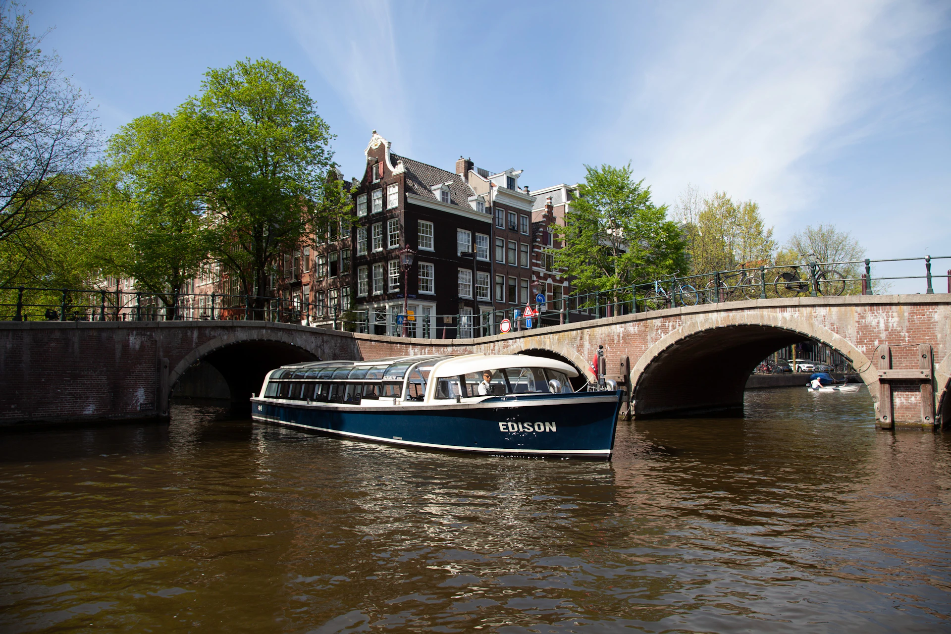 A boat named Edison floating on a canal in front of a bridge and traditional Dutch-style buildings under a clear sky.