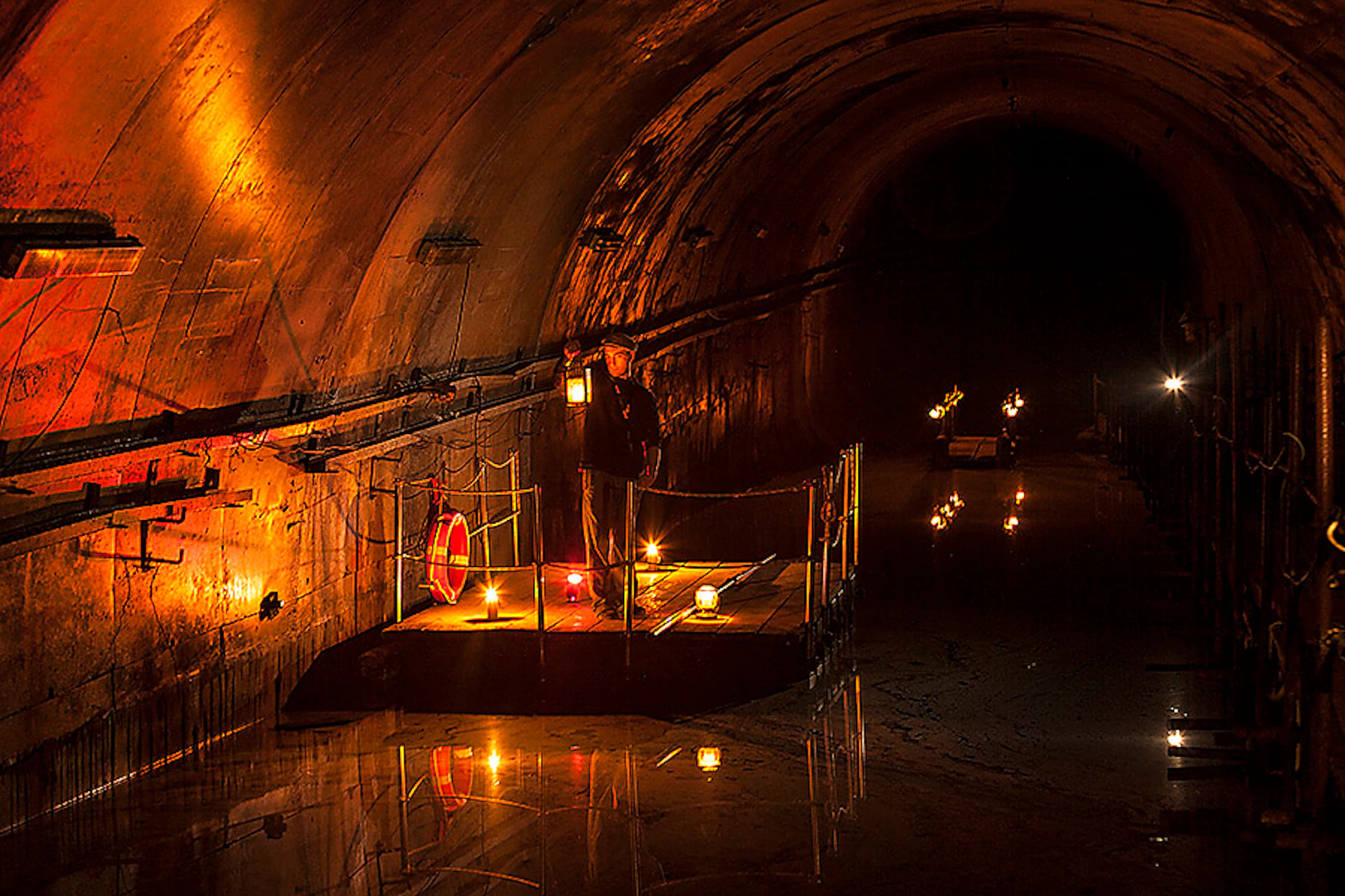 A person holding a lantern stands on a small, illuminated platform inside a dark, arched tunnel reflected in water.