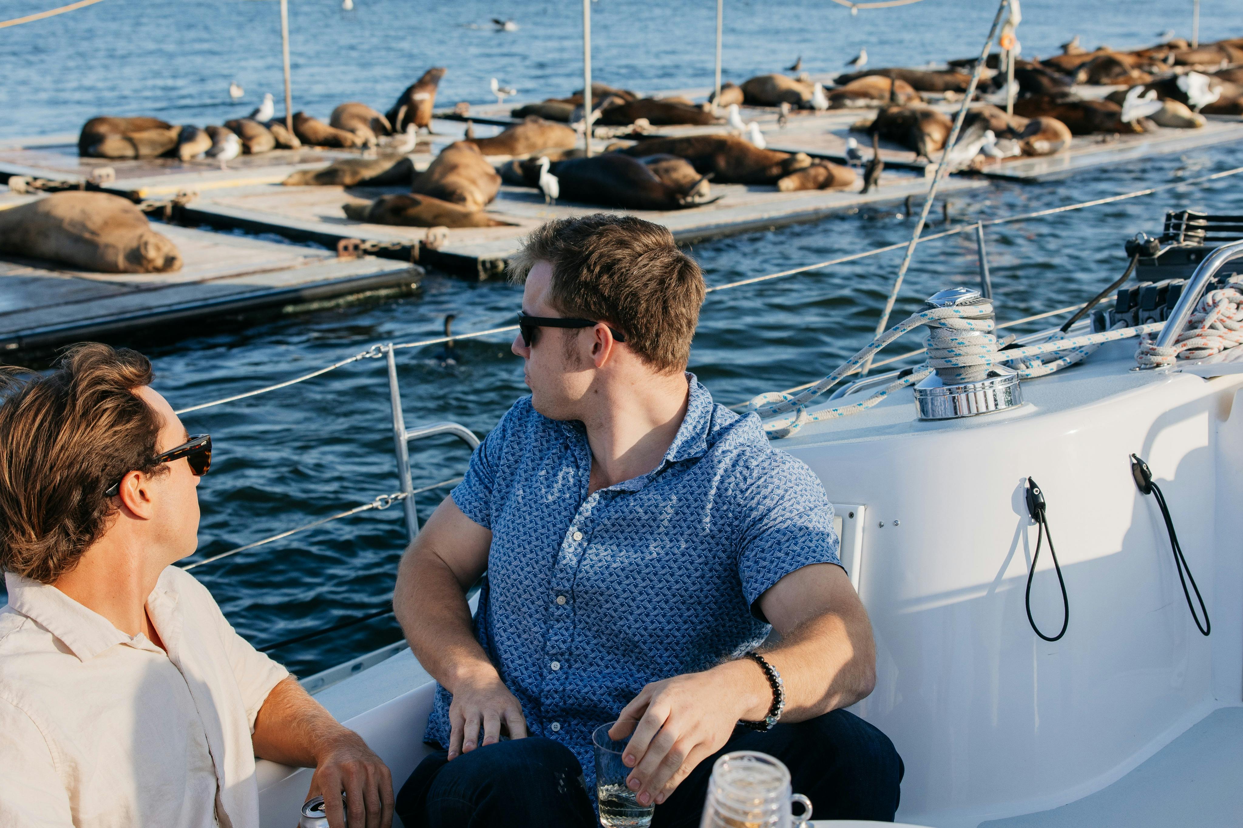 Two young males sitting on a sailboat turning to watch the sea lions lounging behind them