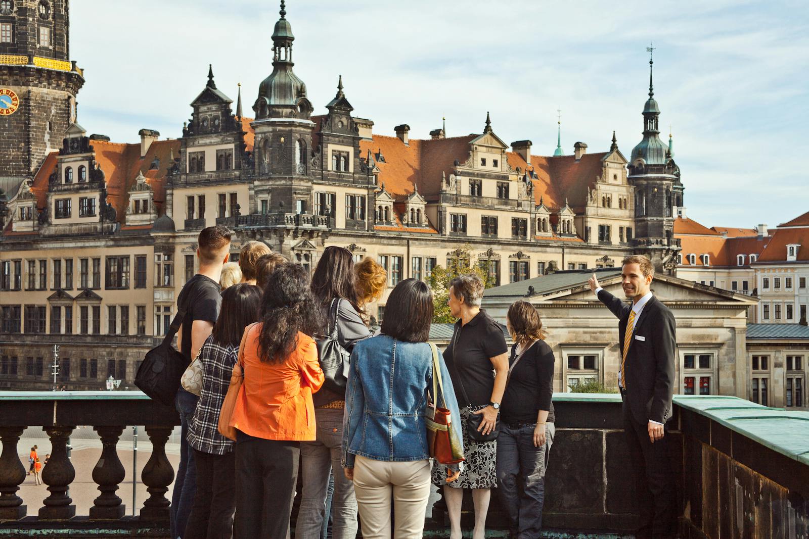 A tour guide points at an ornate historical building as a group of people attentively listen and look in the same direction.