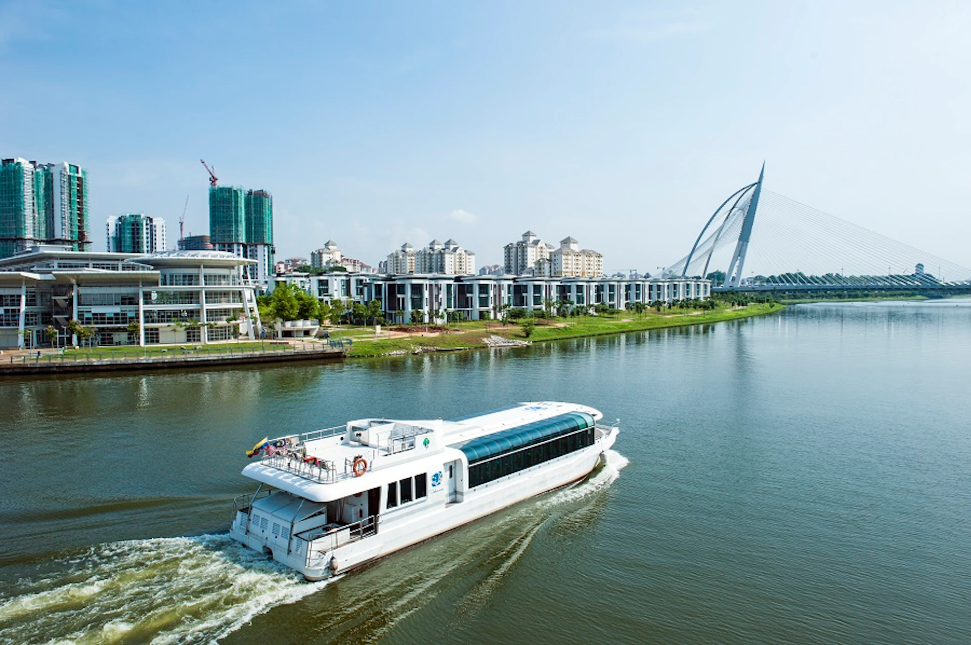 A white passenger boat navigates a river alongside modern buildings and a distinctive cable-stayed bridge on a clear day.