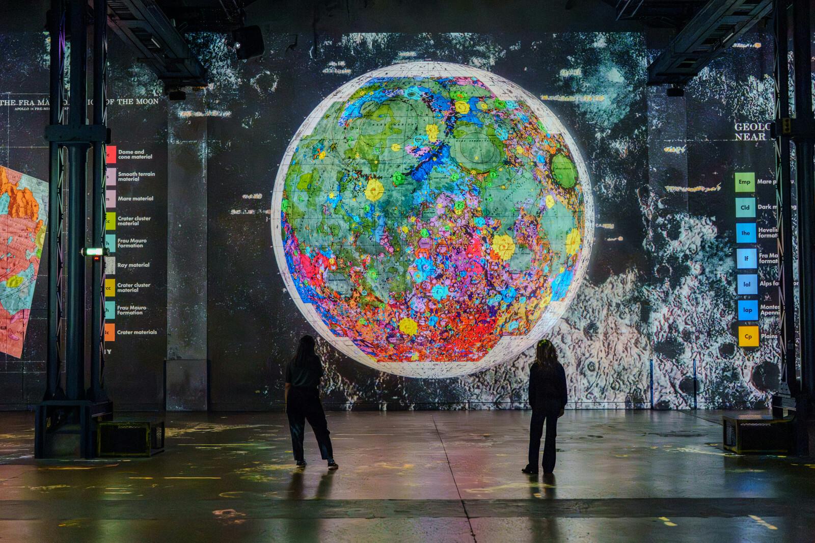 Two people stand facing a large, colorful digital display of a detailed moon map projected onto a wall in a dimly lit room.