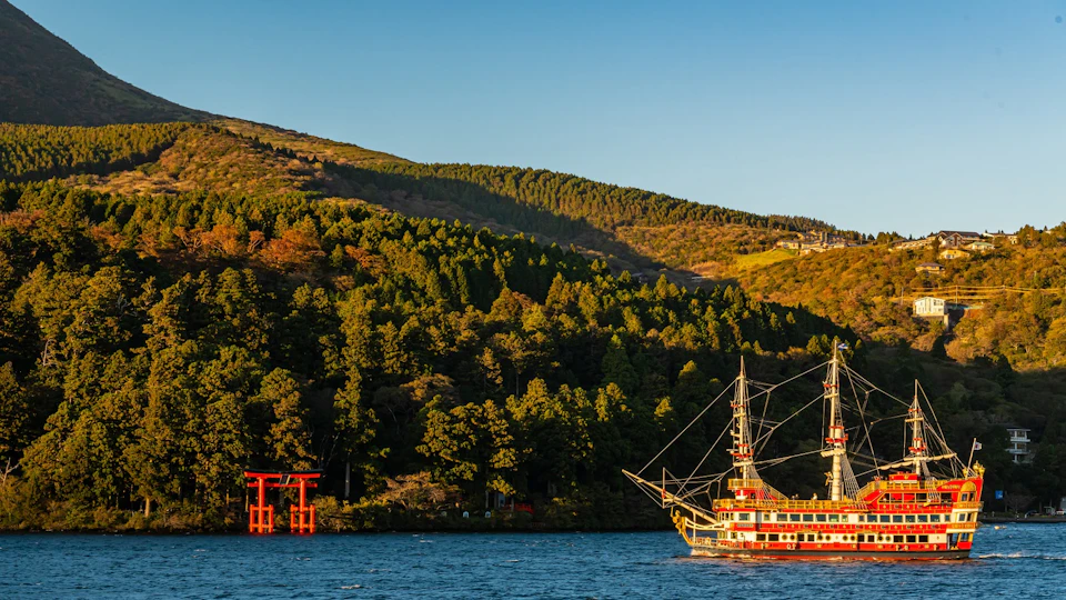 A scenic view of a lake with a red pirate-style ship and a red torii gate, surrounded by forested hills under clear skies.