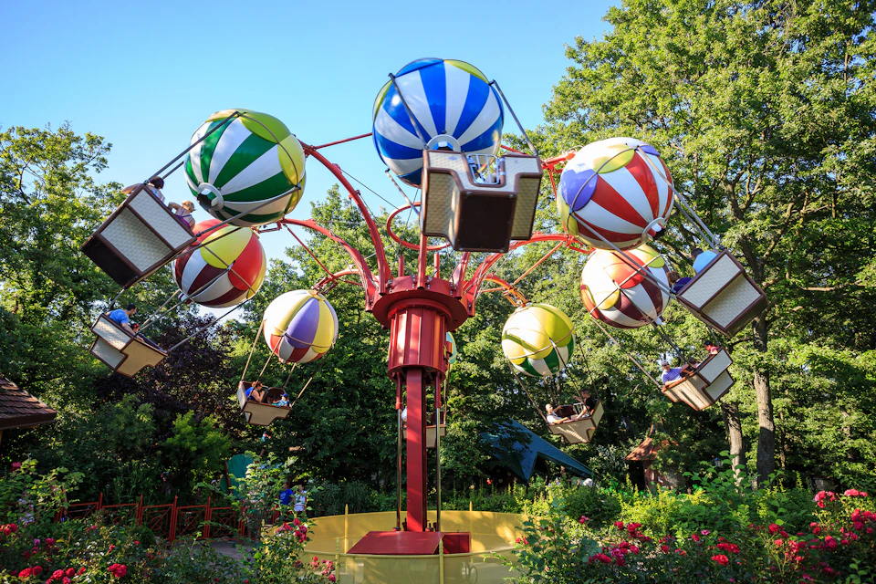 Colorful balloon-themed amusement ride with seated passengers, set against a backdrop of lush greenery and a clear blue sky.