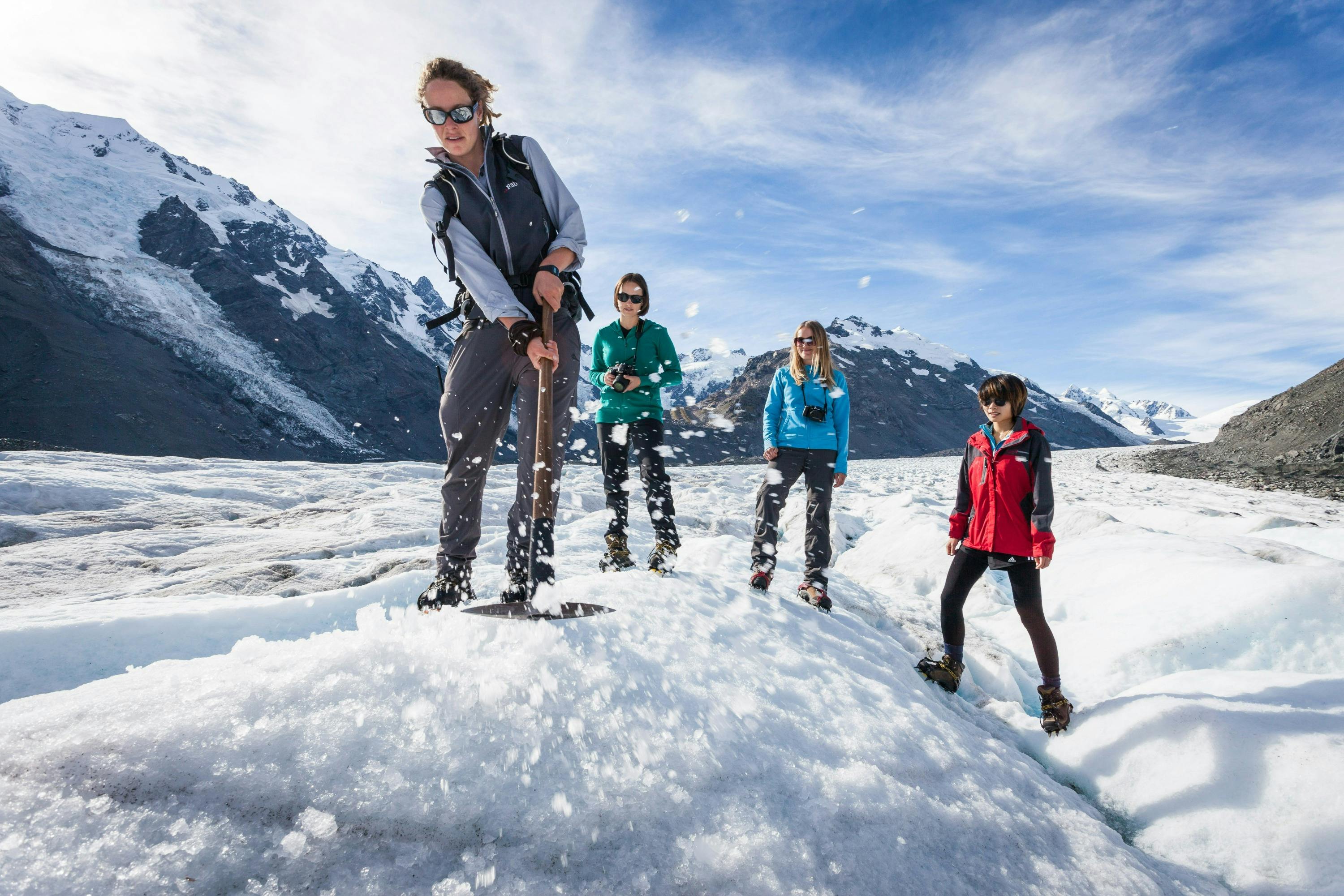 Four people stand on a snowy glacier, one digging with a pickaxe, under a bright blue sky with mountains in the background.