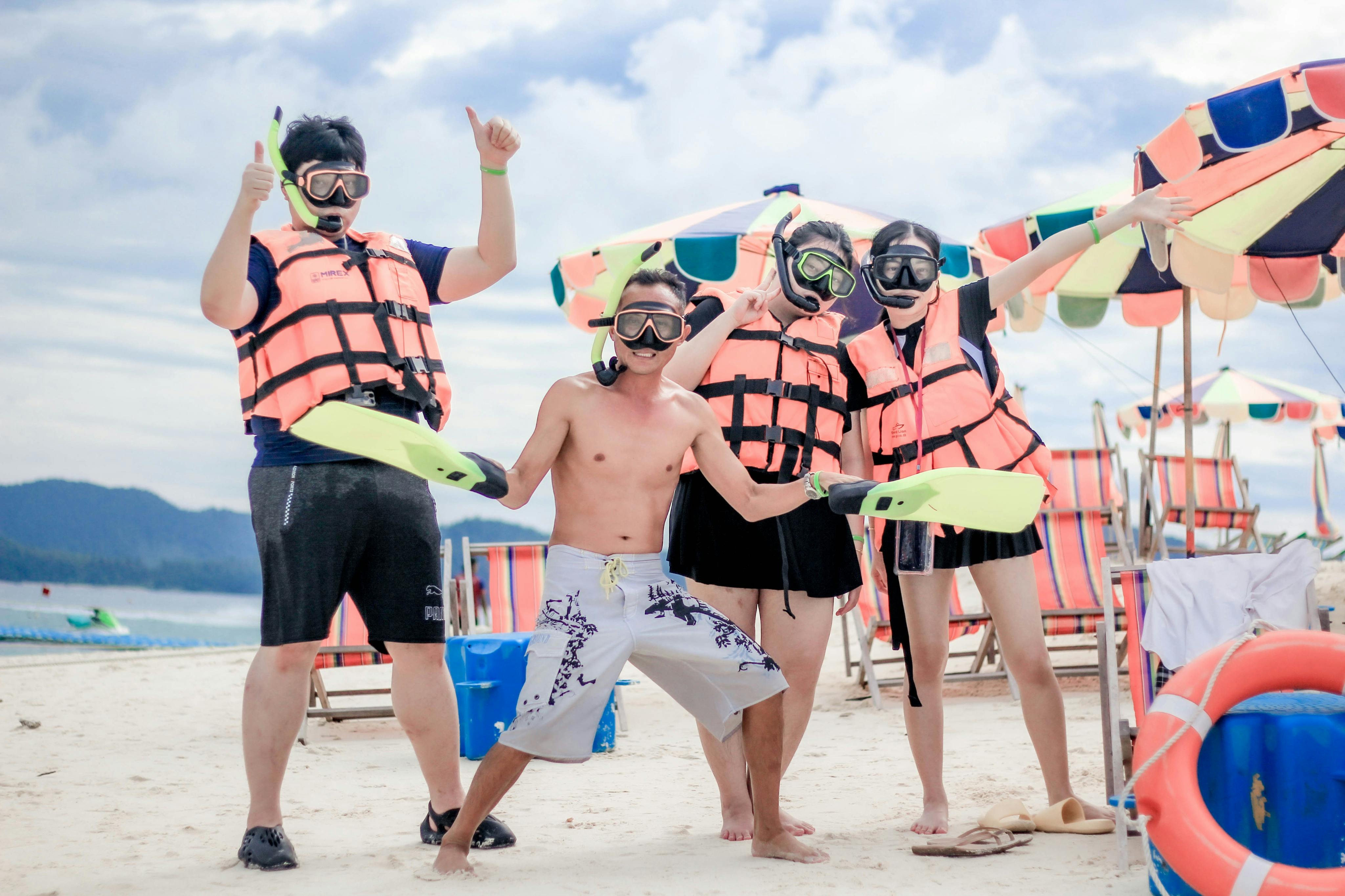 Four people in snorkeling gear, including life jackets and flippers, pose excitedly on a beach with colorful umbrellas.