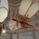 Una macchina volante vintage con grandi ali in tessuto e ingranaggi in legno, sospesa al chiuso con un soffitto intricato sullo sfondo.