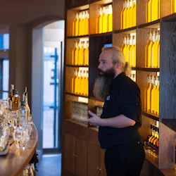 En skäggig man gestikulerar och talar till en grupp människor i en bar kantad med flaskor med gul vätska.