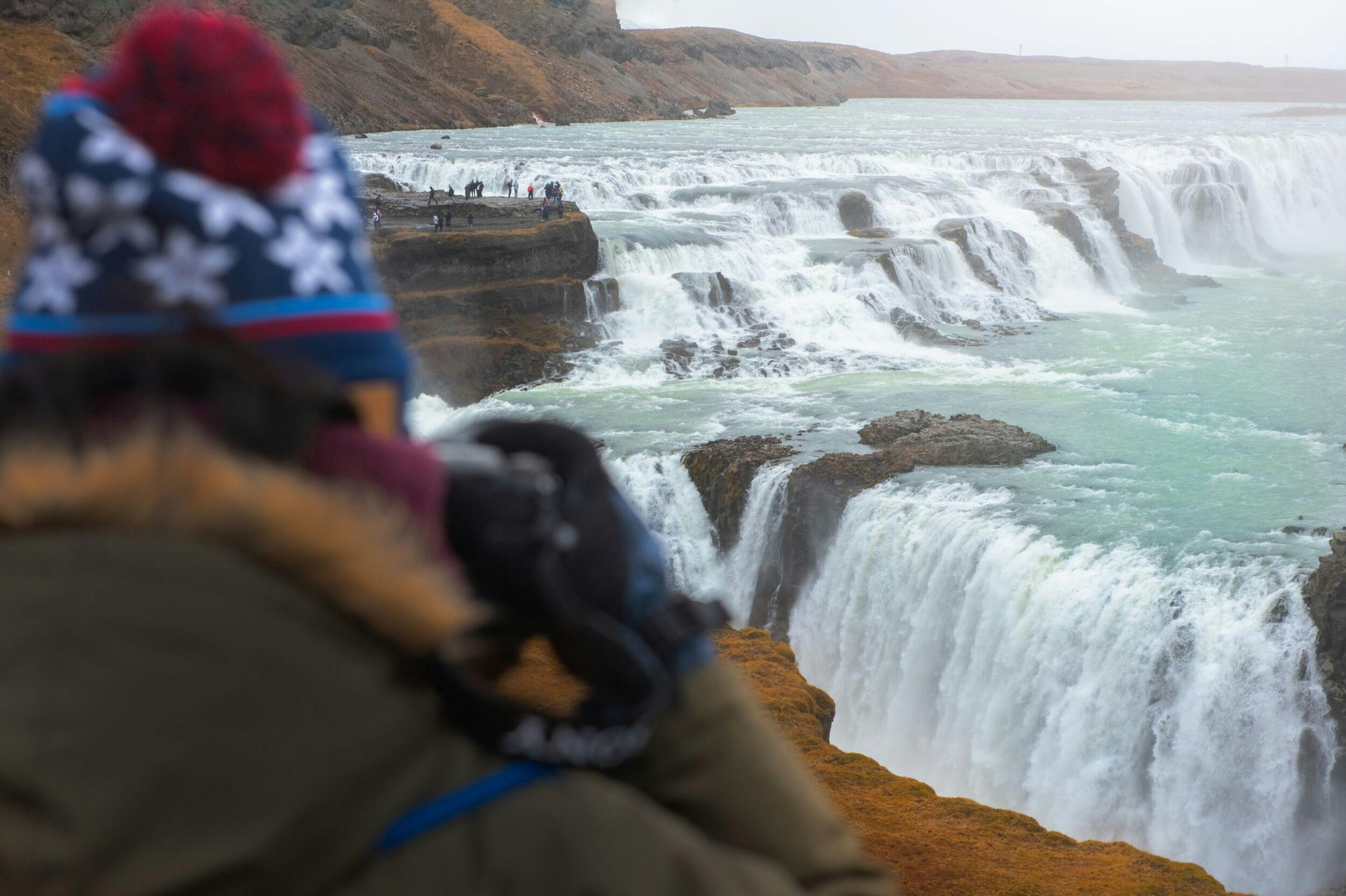 A person in a winter hat observes a large, multi-tiered waterfall surrounded by rocky terrain and distant spectators.