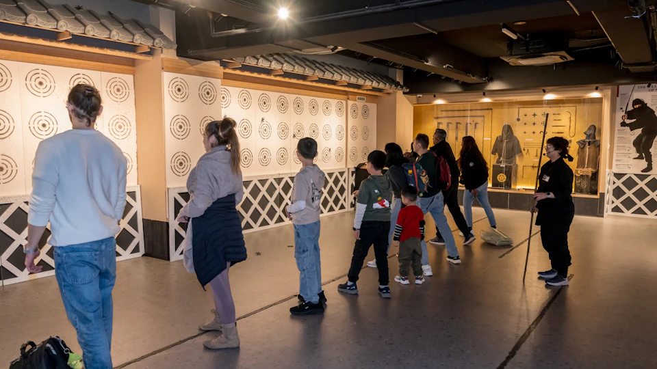 People standing in line at an indoor archery or shooting range with target boards displayed on the wall.