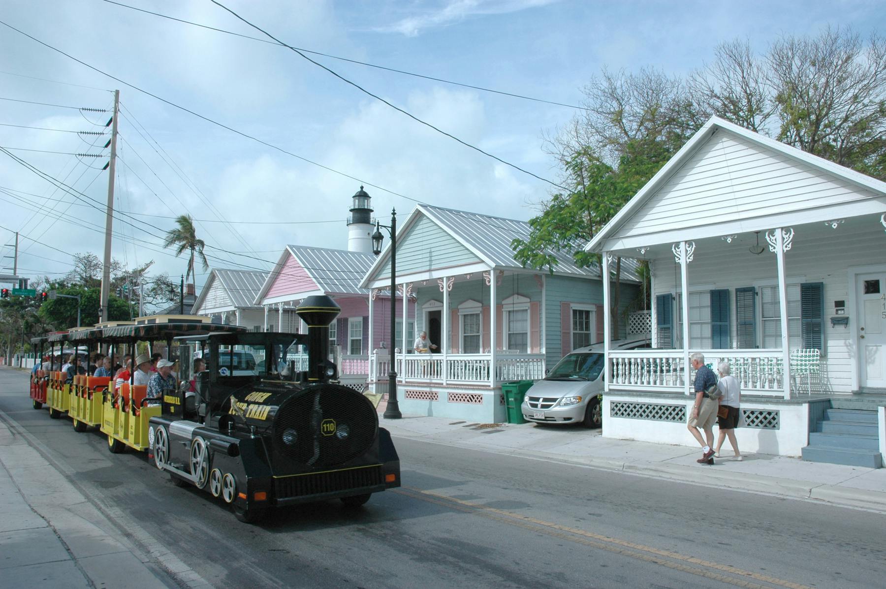 Un petit train touristique noir circule dans une rue bordée de maisons aux couleurs pastel, avec un phare en arrière-plan.