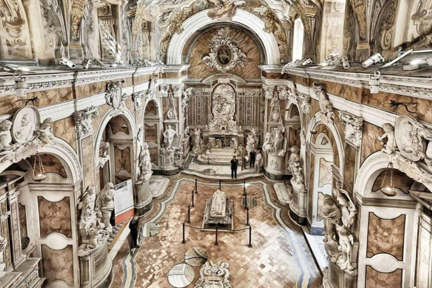 Una sala ornamentada amb escultures, arcs i una decoració intrincada, amb una tomba de pedra central i visitants observant l'espai.