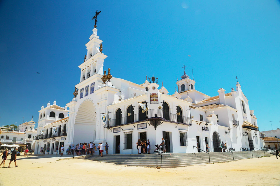 White building with ornate architecture and bell tower under clear blue sky. Groups of people gather at the entrance and steps.