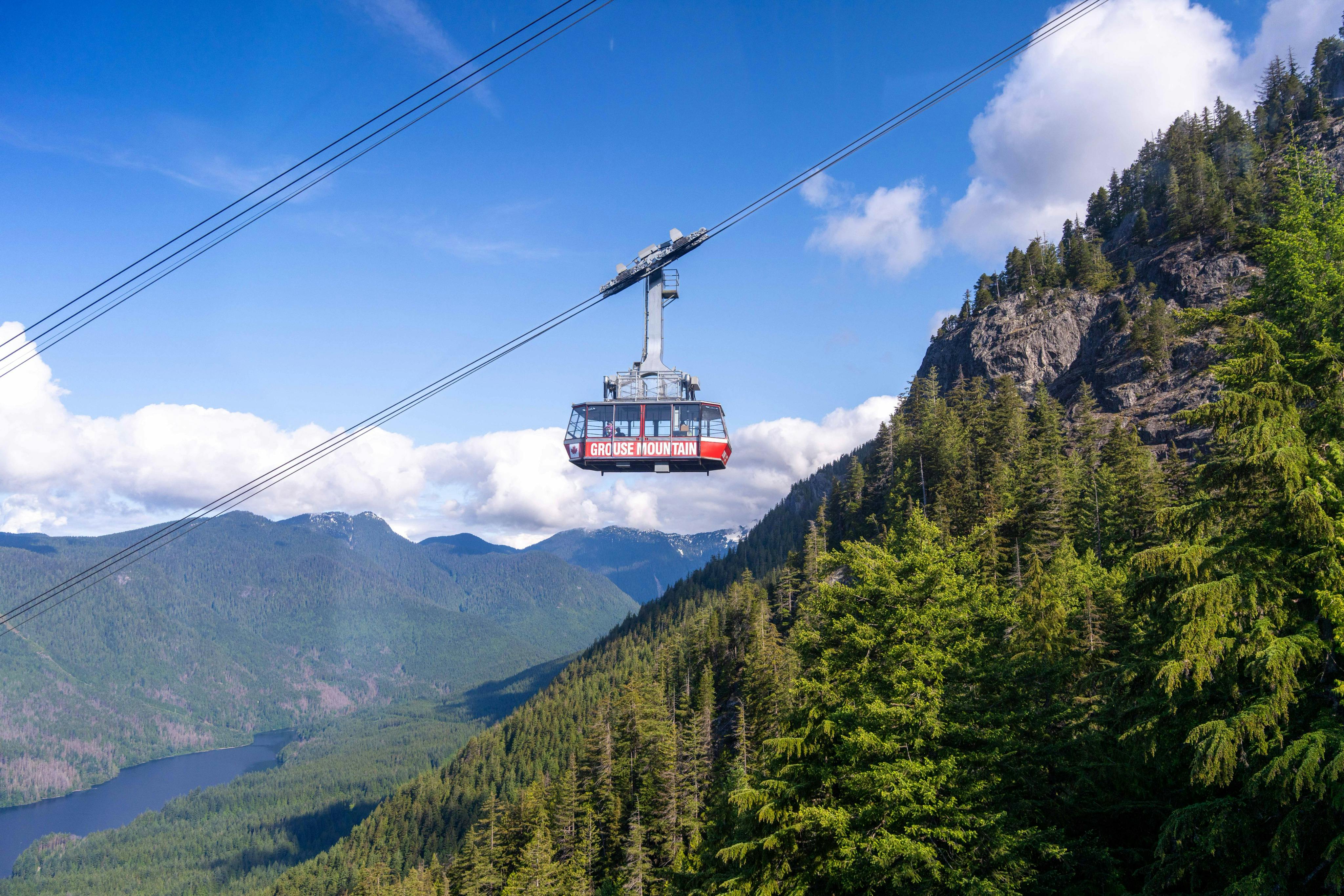 Nad zalesionym krajobrazem górskim pod błękitnym niebem zawieszona jest czerwona kolejka linowa z napisem „Grouse Mountain”.