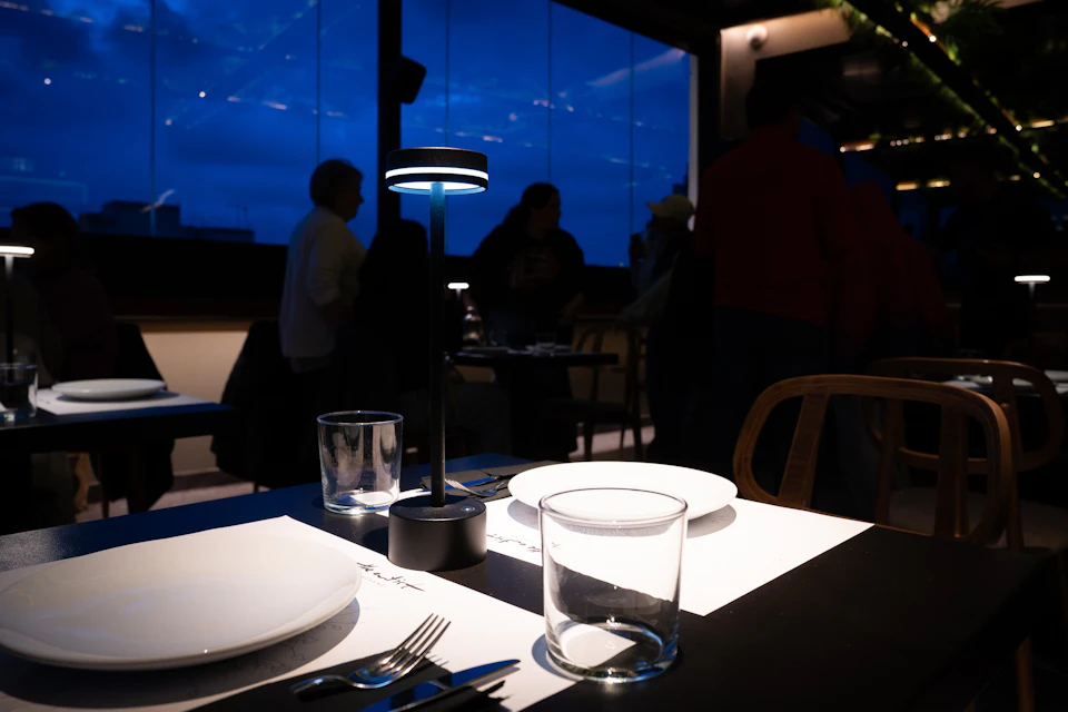 A dining table setup with plates, glasses, and cutlery under a small lamp, with people in a dimly lit background.