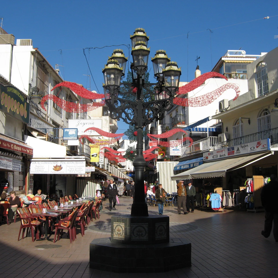 Outdoor market with shops, a decorated street lamp, dining tables with chairs on the left, and people walking and shopping.