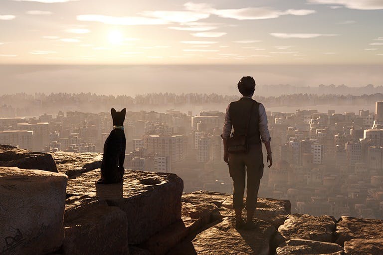 Una persona i un gat negre s'aixequen en un aflorament rocós amb vistes a un paisatge urbà sota un cel ennuvolat durant la posta de sol.