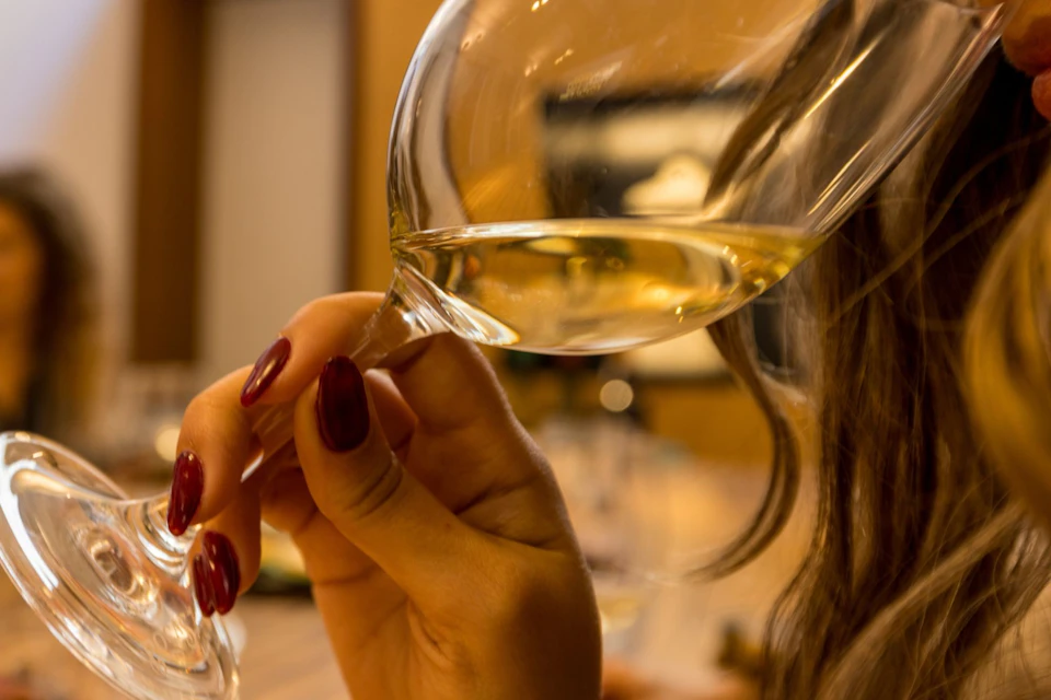 Close-up of a hand holding a wine glass with white wine, with blurred background and warm indoor lighting.