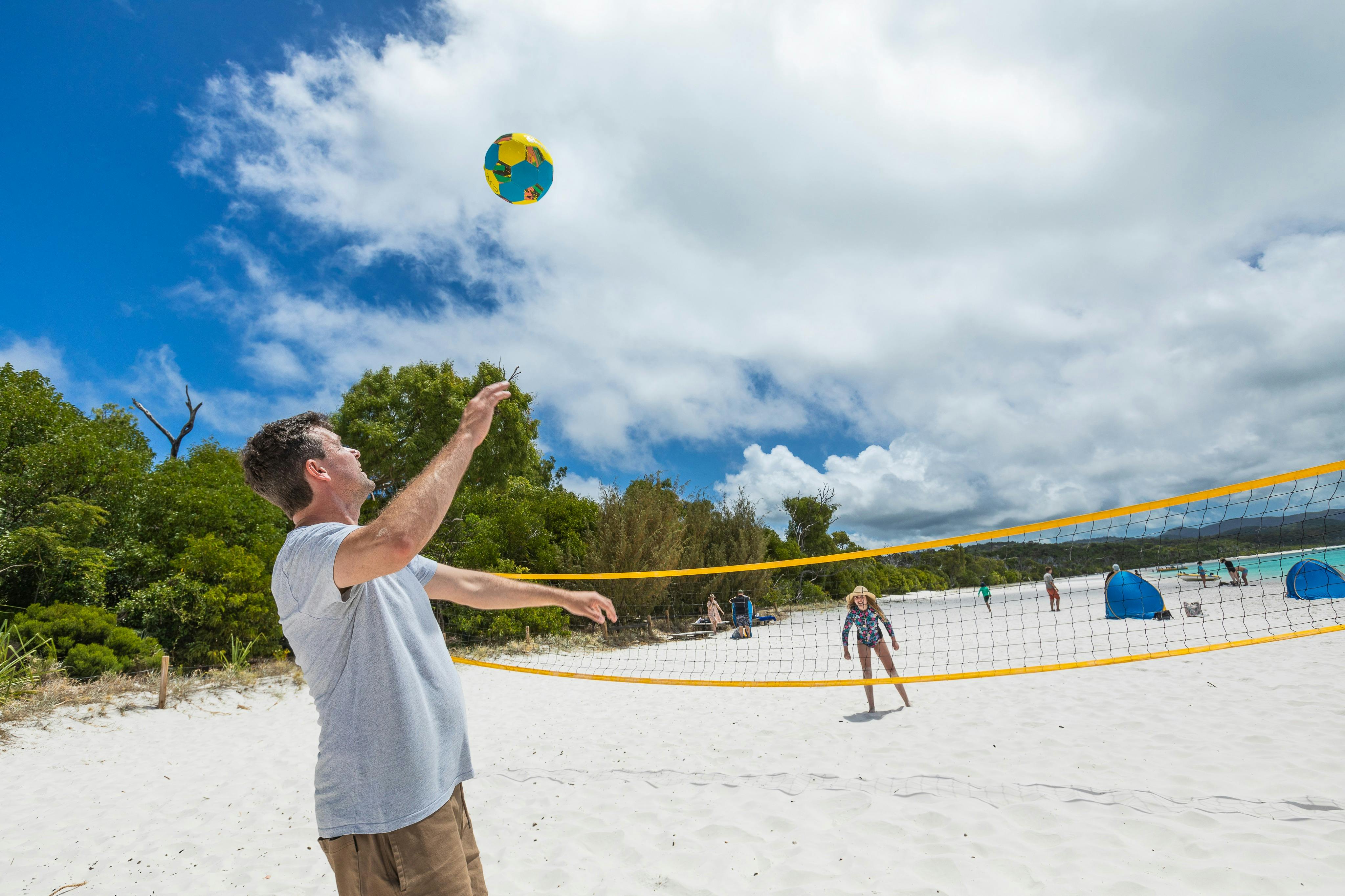 Persone che giocano a beach volley in una giornata di sole, con alberi e sabbia bianca sullo sfondo. Uomo in camicia grigia che colpisce la palla.