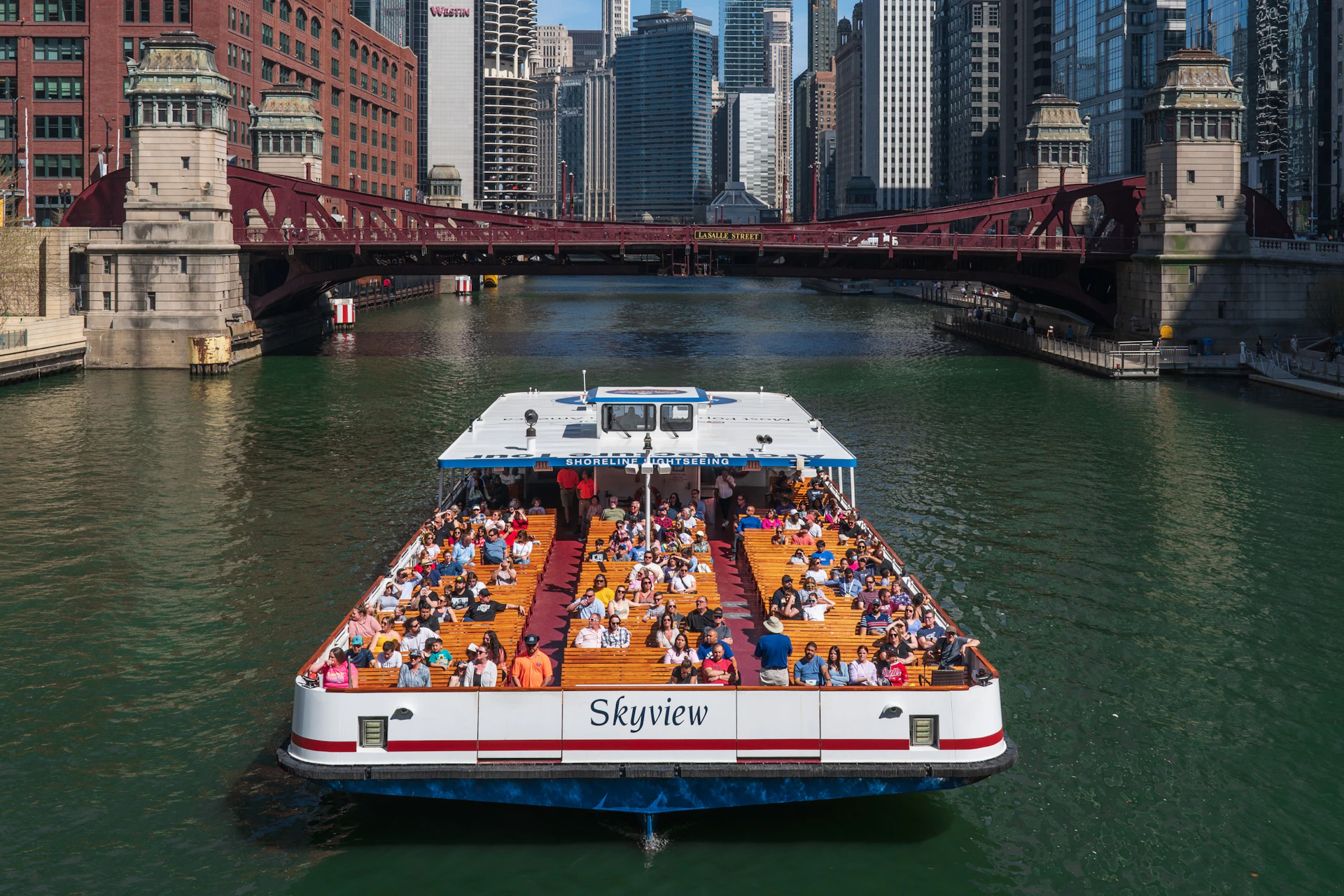 A sightseeing boat named 'Skyview' filled with people cruising on a river through a city with tall buildings and a bridge.