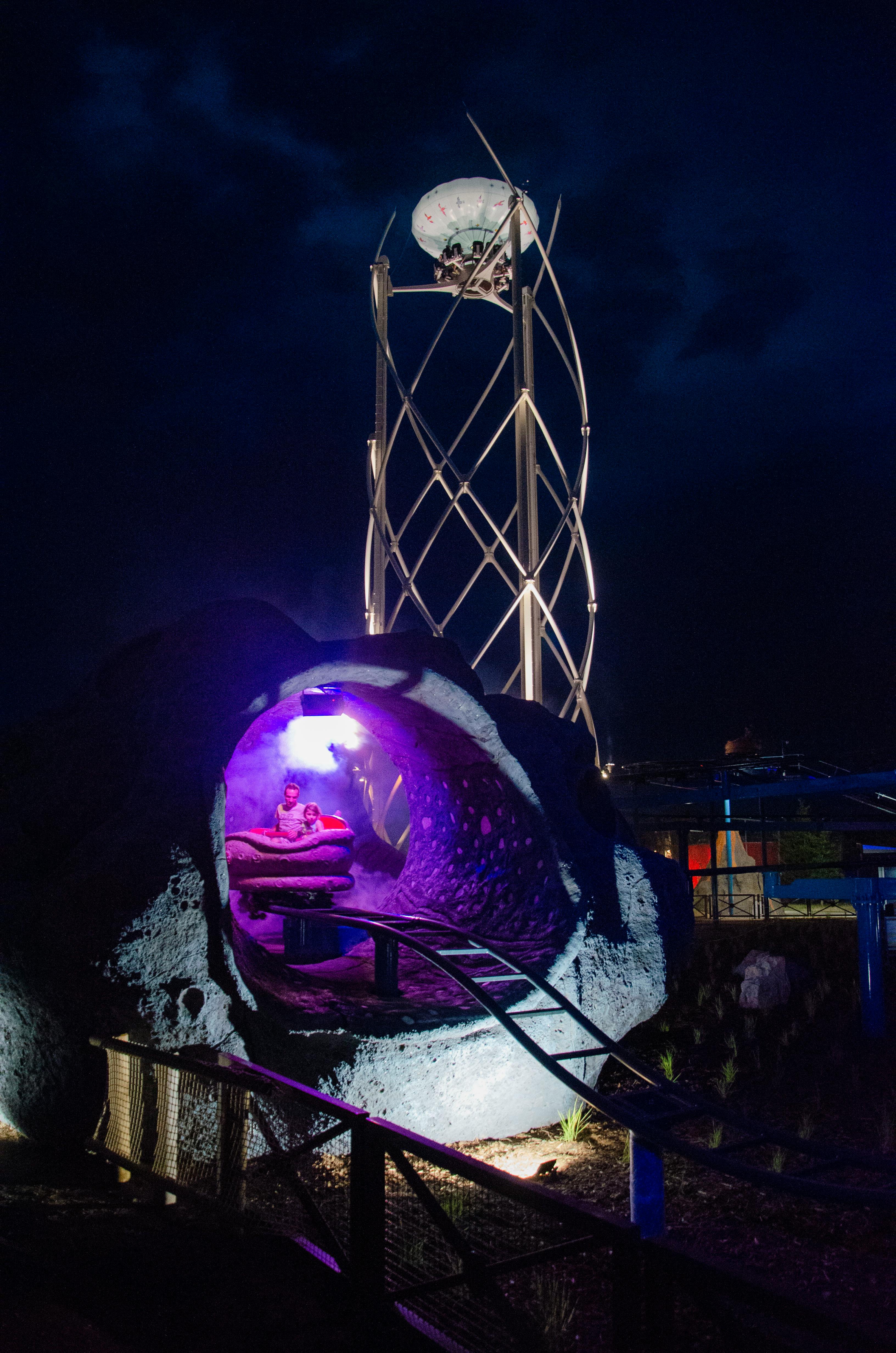 두 명의 라이더가 탑승한 보라색 롤러코스터 터널, 높은 조명 구조물, 어두운 밤하늘을 배경으로 펼쳐집니다.