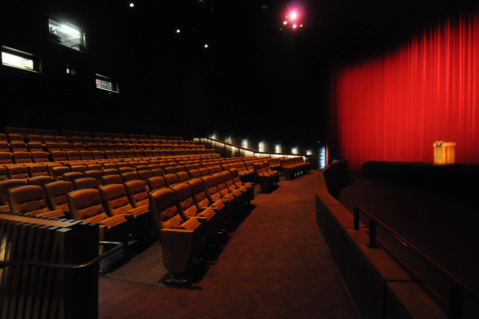 An empty theater with rows of seats, a red curtain on the stage, and dim lighting.