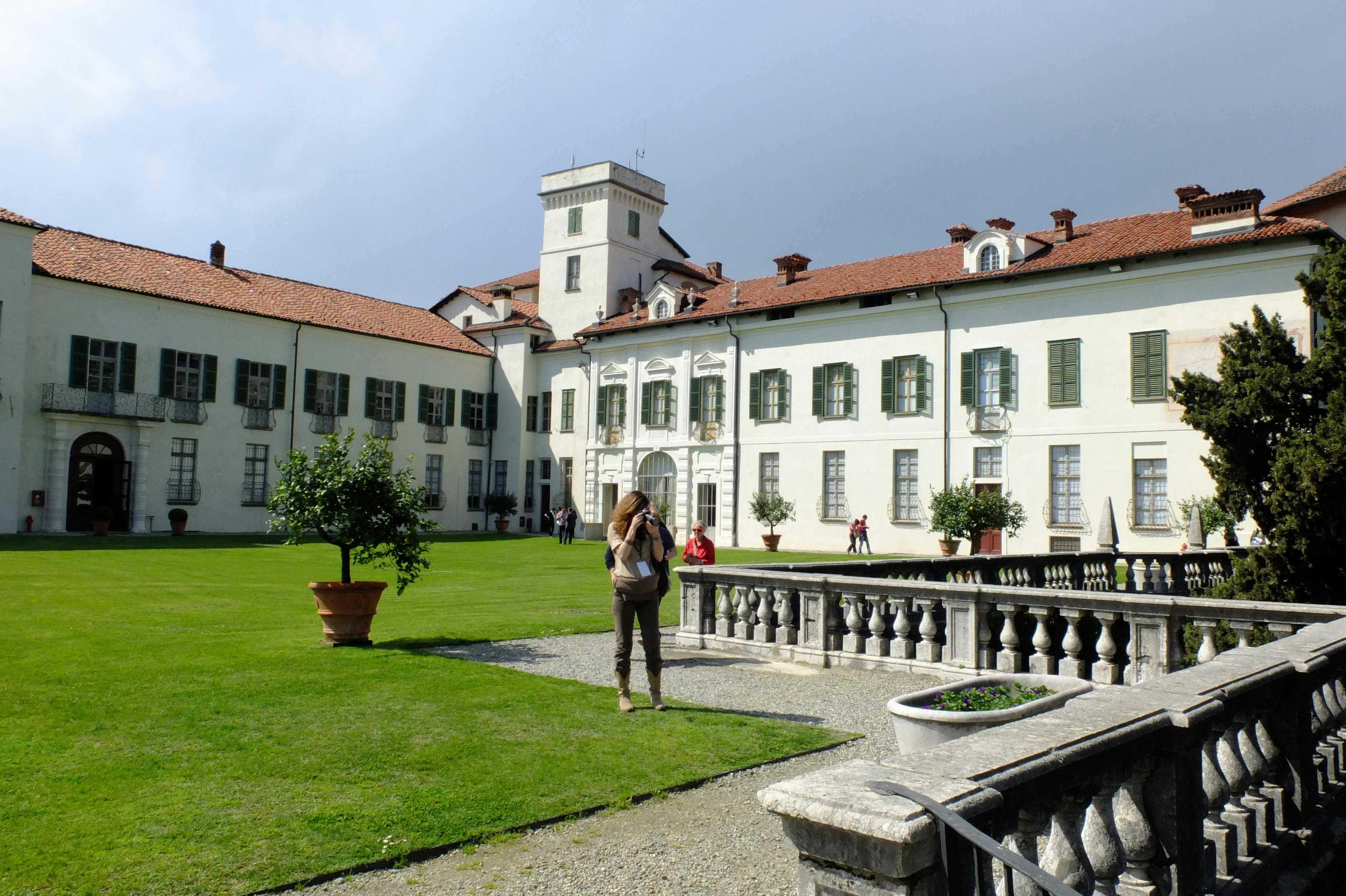 A person stands in a courtyard photographing an old, white building with green shutters and a red-tiled roof under a cloudy sky.