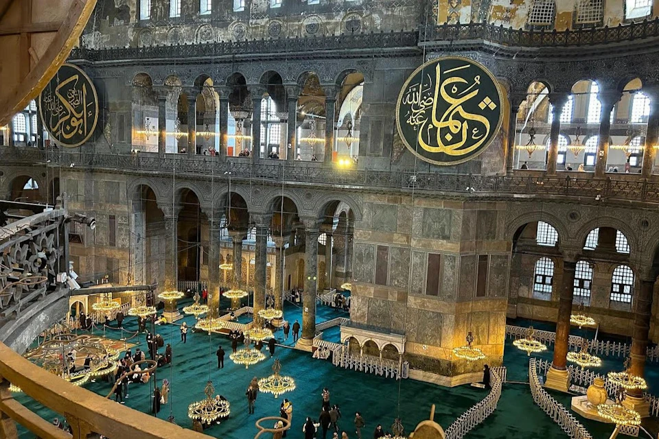 Interior of the Hagia Sophia, featuring ornate chandeliers, Islamic calligraphy, and visitors on a green carpet.