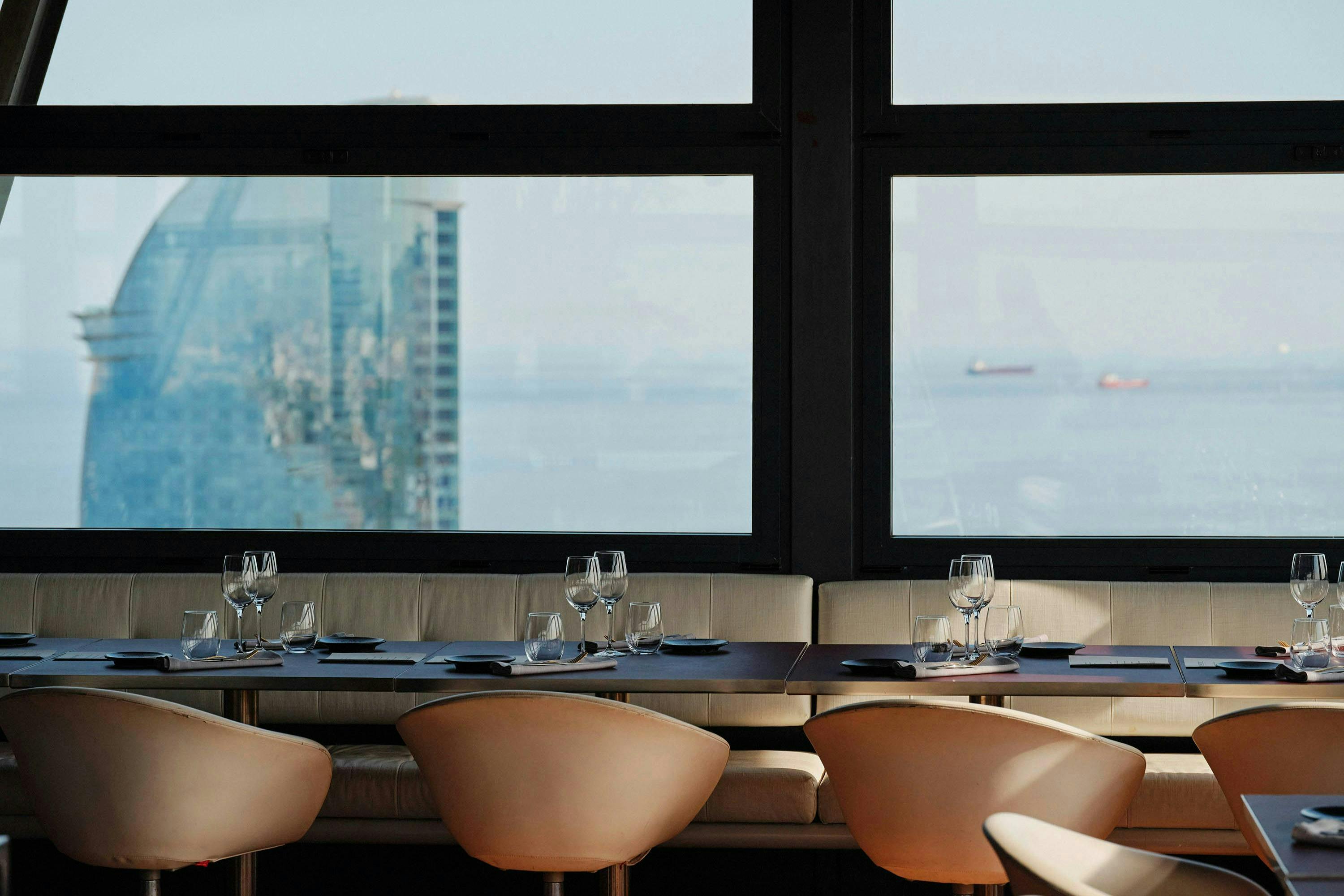 Modern restauranginredning med dukade bord och stolar, stort fönster som visar en stadsbild och fartyg på vattnet i bakgrunden.