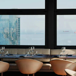 Modern restauranginredning med dukade bord och stolar, stort fönster som visar en stadsbild och fartyg på vattnet i bakgrunden.