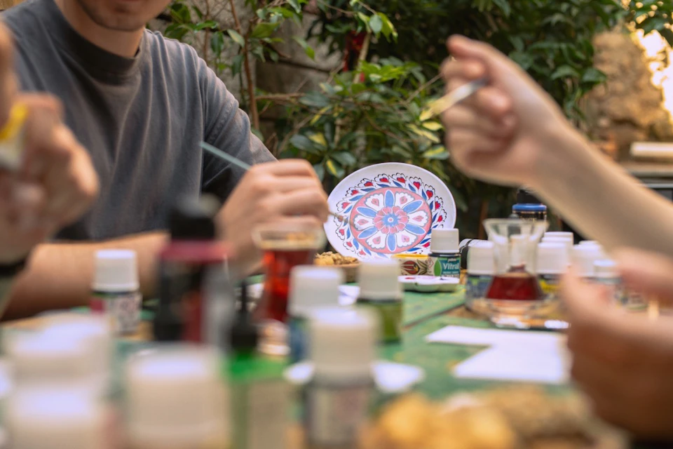 People painting decorative plates on a table outdoors, surrounded by paint bottles and brushes, with greenery in the background.