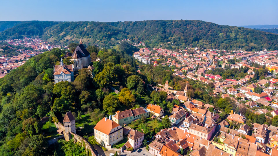 Aerial view of a picturesque village with red-tiled roofs, a large church, lush green trees, and surrounding hills.