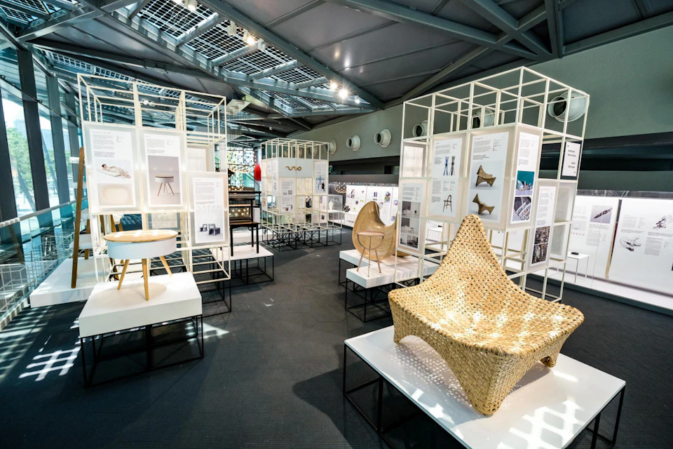 Exhibit showcasing various modern chairs and their designs with informational panels, displayed in a well-lit room with a high ceiling.