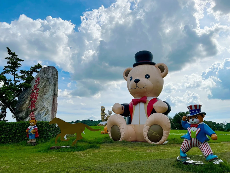 A large inflatable teddy bear wearing a top hat and bow tie, surrounded by smaller statues in a grassy outdoor area.