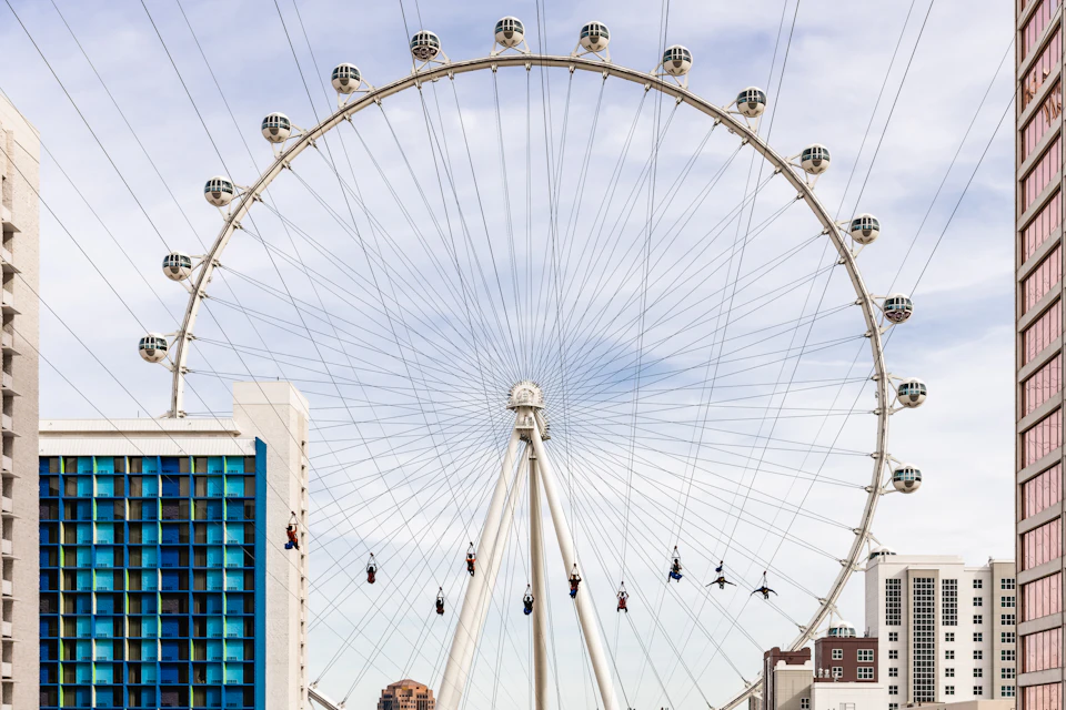 A large Ferris wheel with passenger pods towers over buildings and a zip line with people suspended in mid-air.
