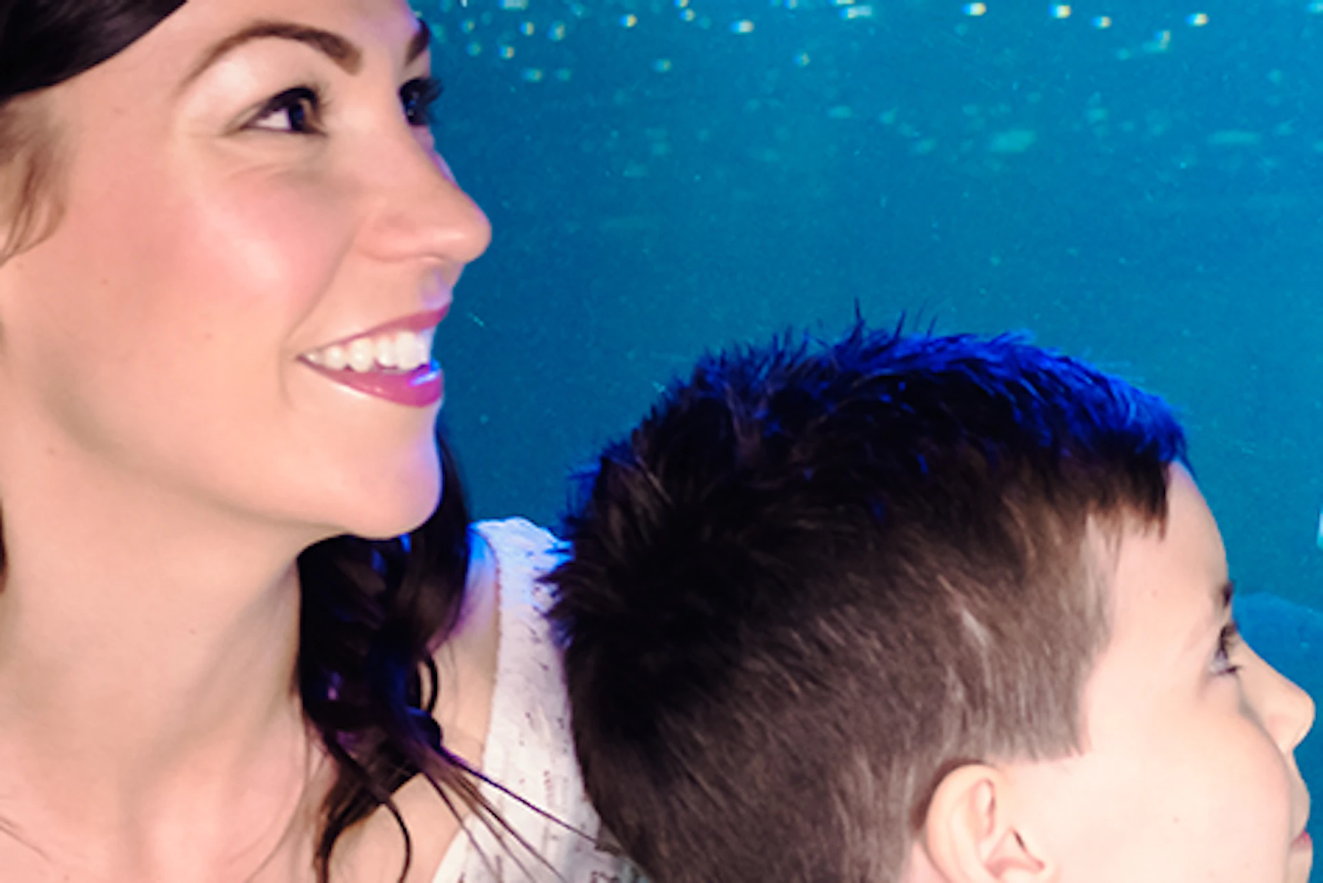 A woman and boy watch a stingray swimming in an aquarium tank. The boy leans on the railing, and colorful fish are visible in the background.