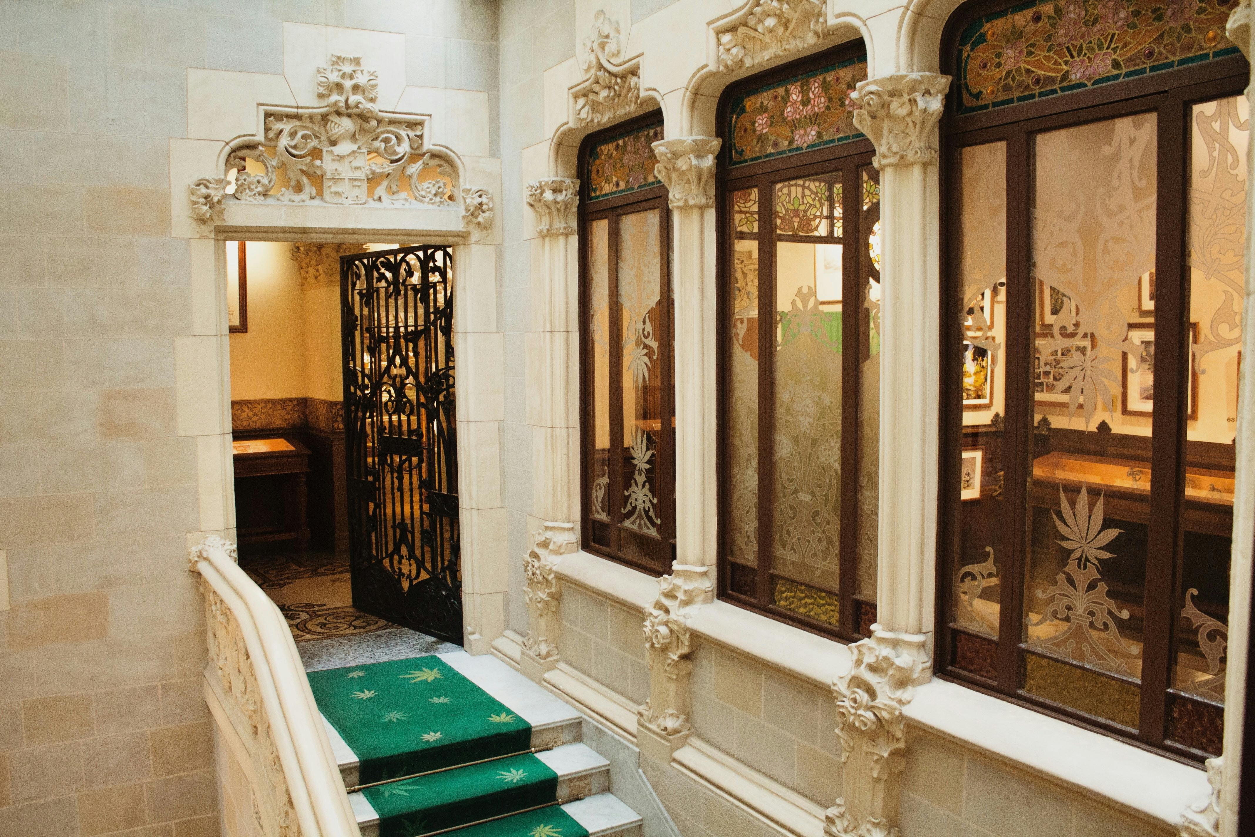 Ornate hallway with decorative arches, stained glass windows, green carpeted stairs, and an intricate iron gate.