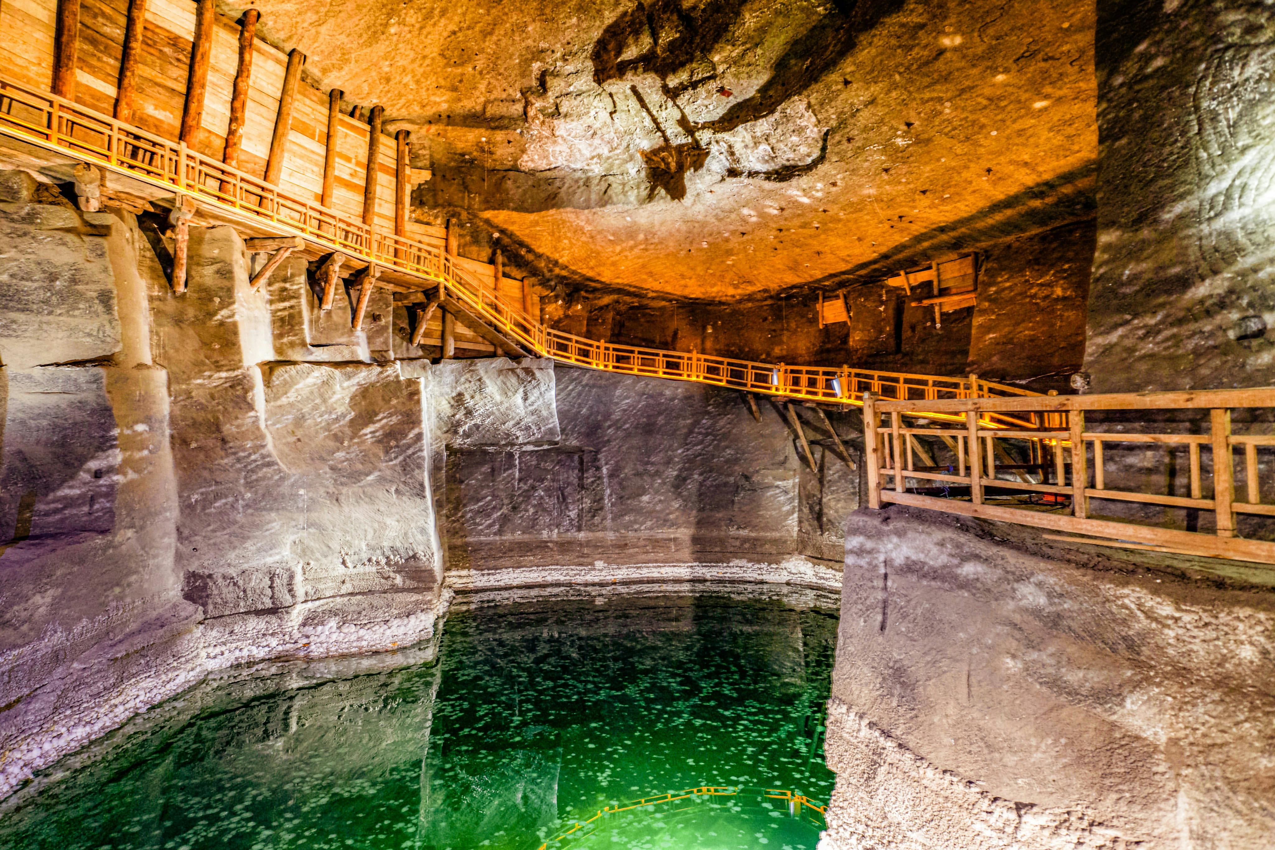 Wooden walkways and stairs inside a large underground cave with a pool of green water below and salt formations visible on the walls.