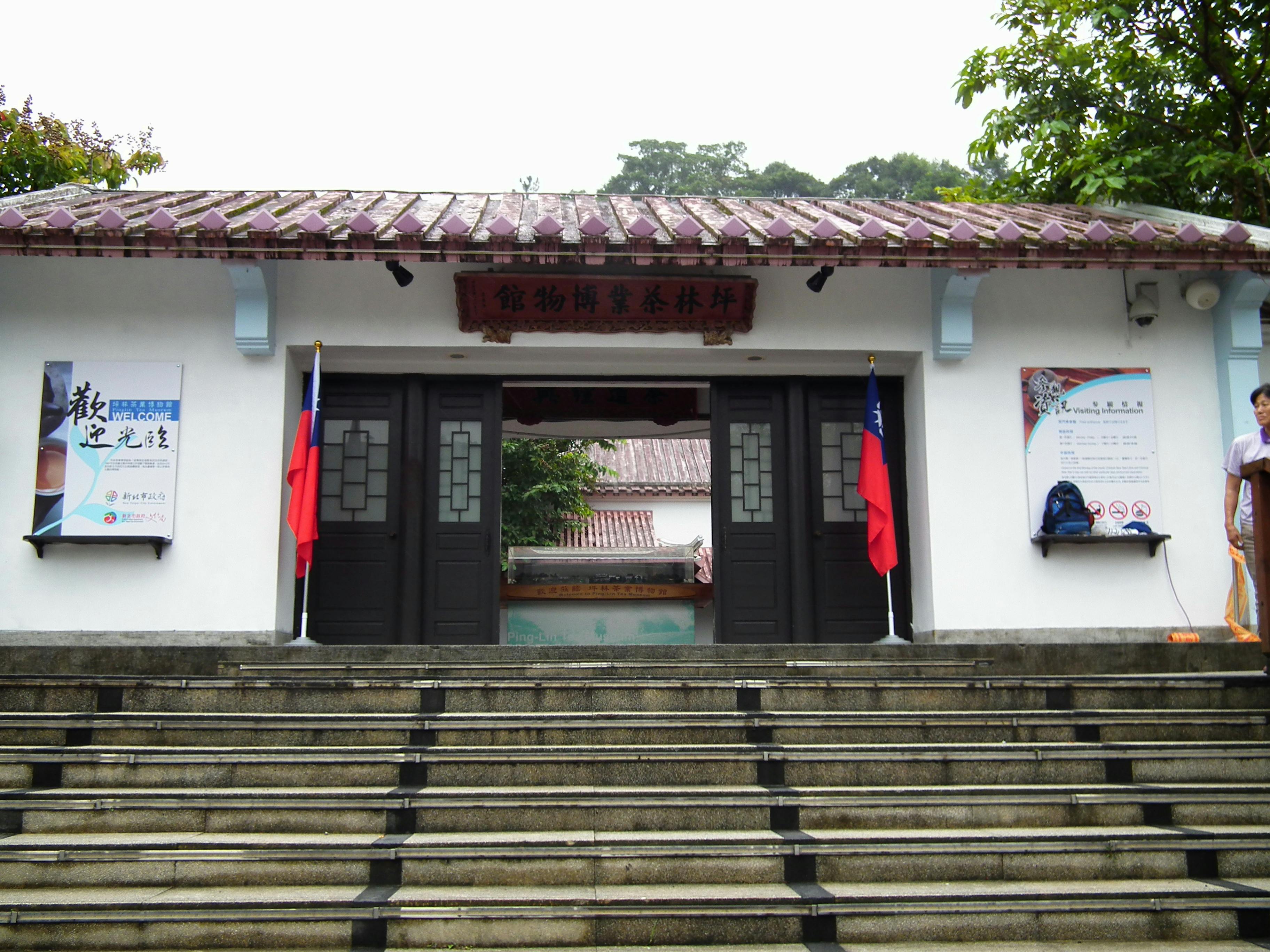 Scalinata in pietra che conduce all'ingresso di un edificio tradizionale con doppie porte in legno, affiancate da due bandiere e alberi sullo sfondo.